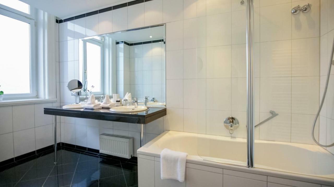 Hotel Ambassador Vienna Luaran gambar The photo shows a modern bathroom featuring a clean, stylish design. There's a large mirror above a double sink vanity area with two sinks and various toiletries arranged neatly. The wall tiles are predominantly white, and the floor is finished in a 